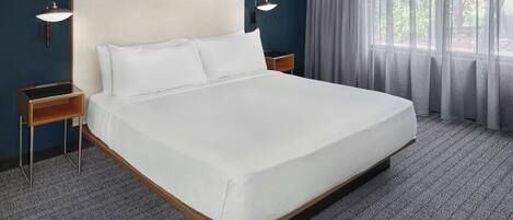 A neatly made bed with white linens in a hotel room. There are two bedside tables with lamps, a window with sheer curtains, and a herringbone-patterned carpet.