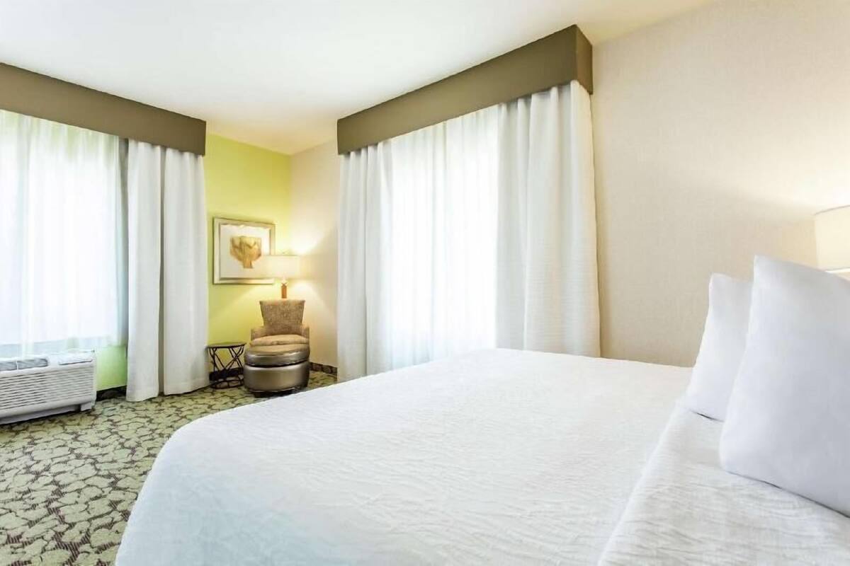 A hotel room with a bed, curtains, a chair, a lamp, and a framed picture on the wall.