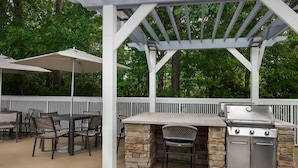 Une terrasse extérieure dotée d’un barbecue en pierre, d’une pergola blanche, ainsi que de chaises et de tables à manger.