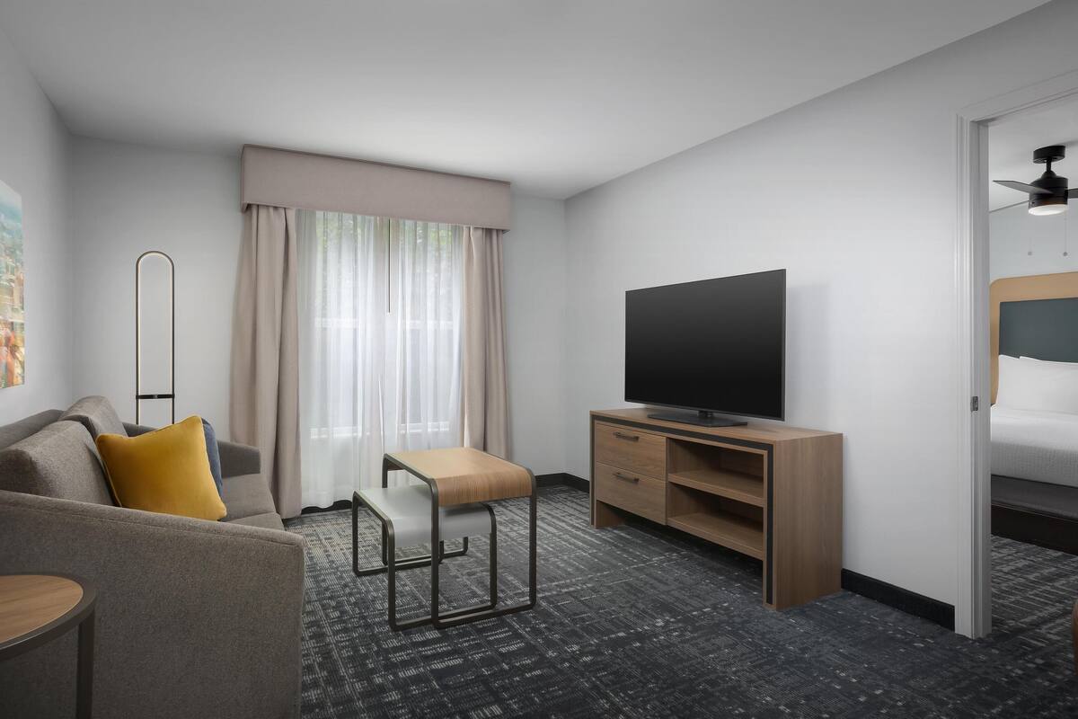Une chambre d’hôtel avec un canapé, une petite table, un téléviseur posé sur un meuble en bois, une fenêtre avec des rideaux et un ventilateur de plafond.