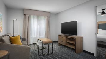 A hotel room with a sofa, a small table, a TV on a wooden stand, a window with curtains, and a ceiling fan.