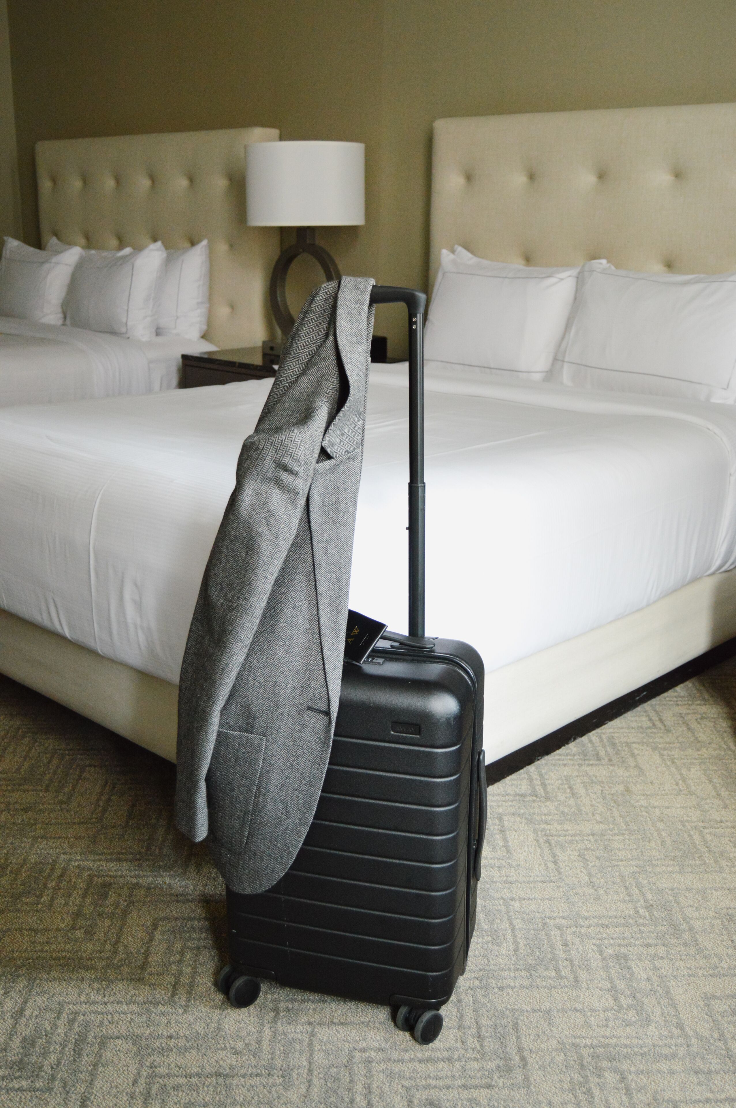 A hotel room with a bed, a lamp, a suitcase, and a jacket on the suitcase.