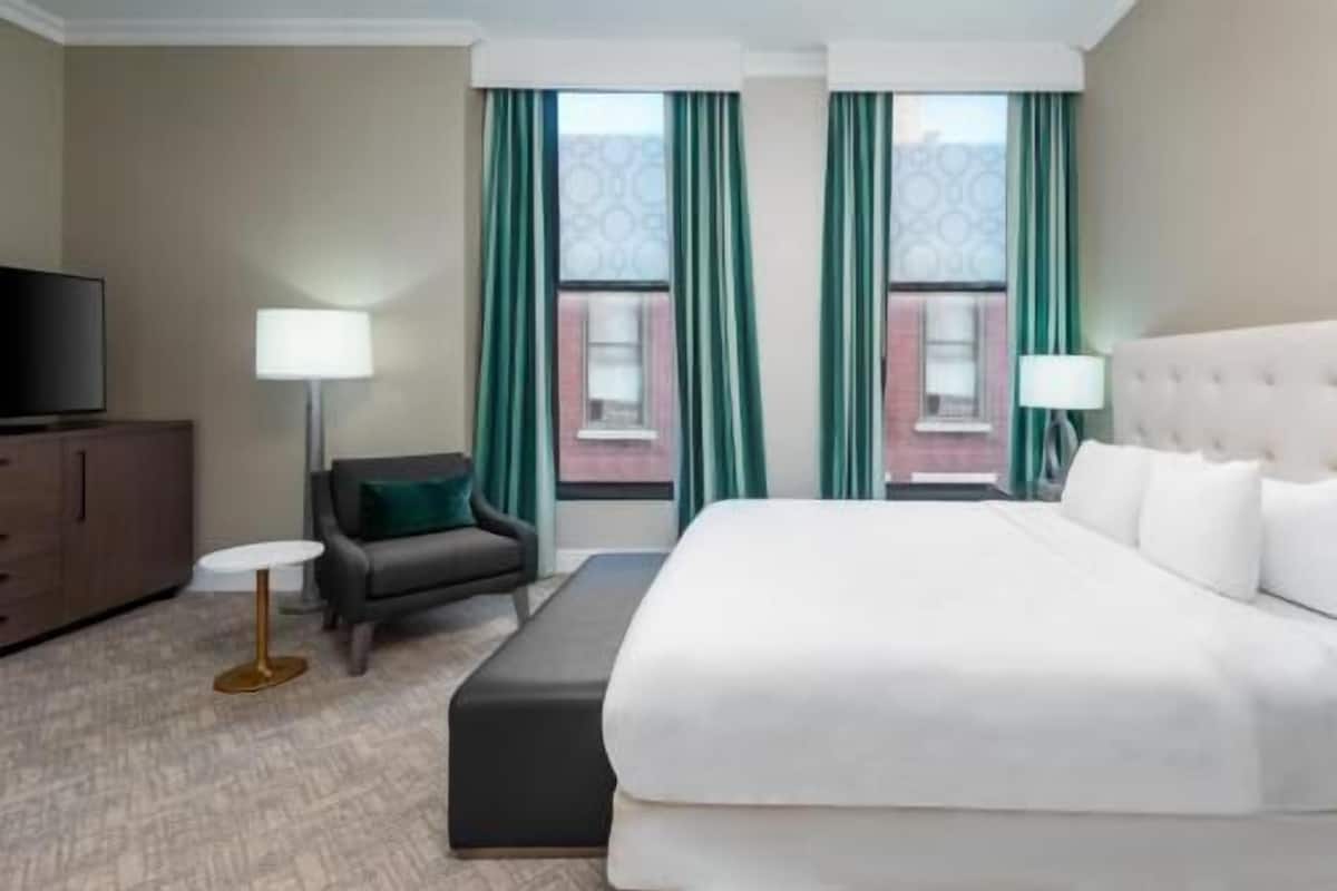 A hotel room with a bed, a TV, a desk, a chair, and two windows with curtains.