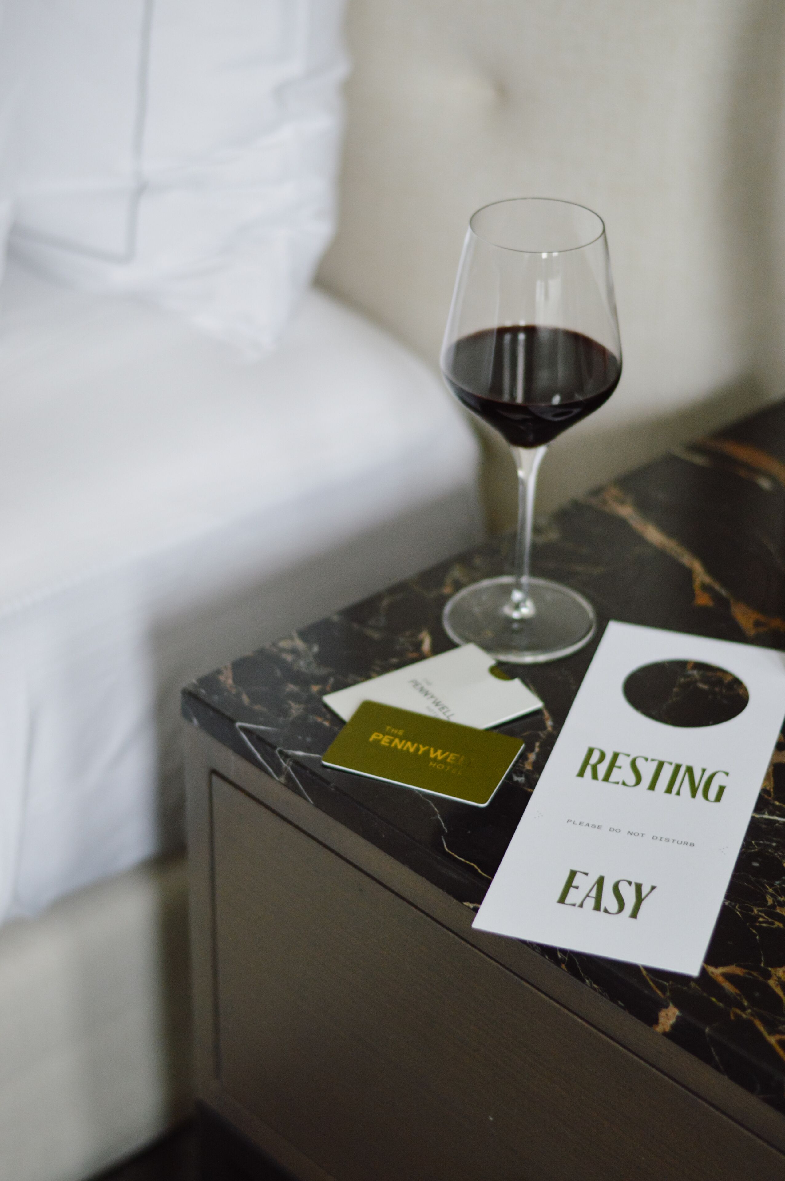 Una copa de vino tinto sobre una mesa de mármol junto a una almohada blanca y un letrero que dice “RESTING EASY”.