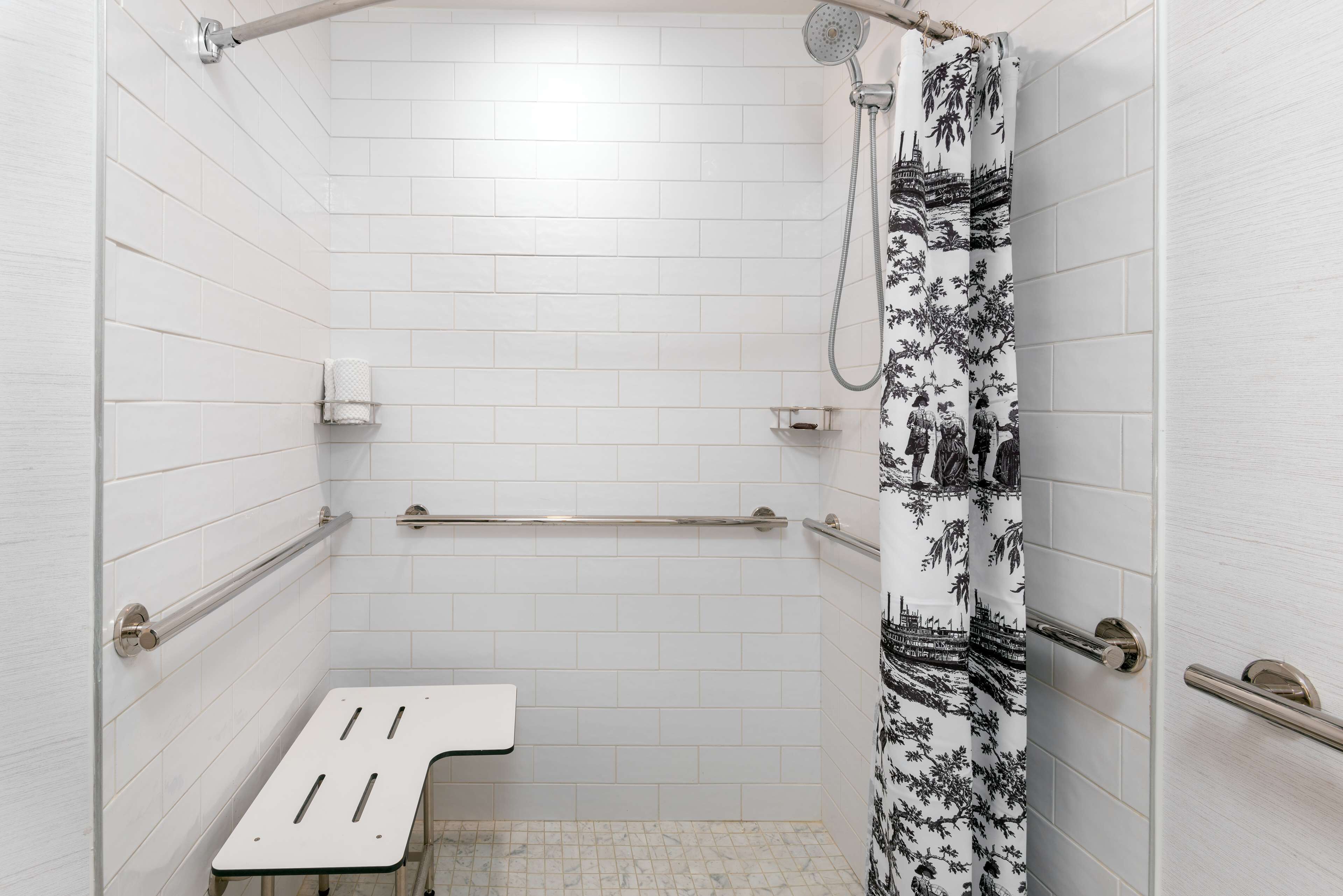 Ein Badezimmer mit weiß gefliesten Wänden, einer Dusche mit schwarz-weiß gemustertem Vorhang, einer Haltestange und einer weißen Sitzbank.