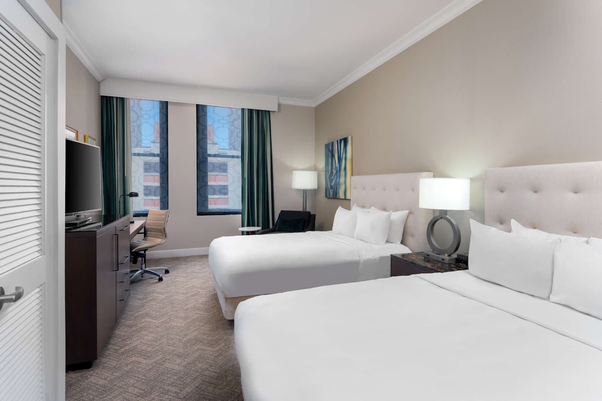 A hotel room with two beds, a desk, a chair, a TV, and a window with curtains.