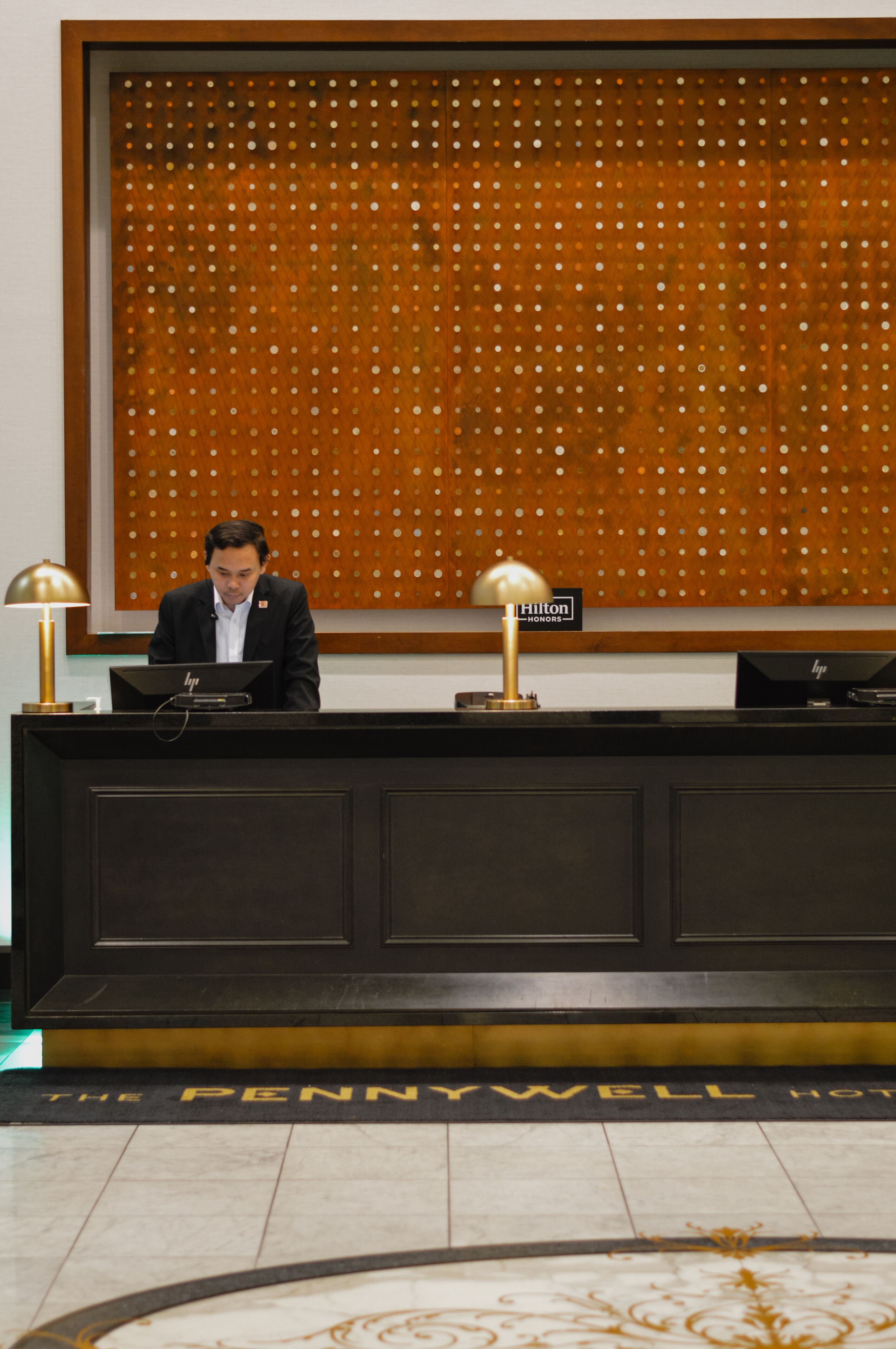 Un comptoir d’accueil d’hôtel avec un homme assis derrière, une enseigne indiquant le programme Hilton Honors et un tapis orné du nom de l’hôtel.