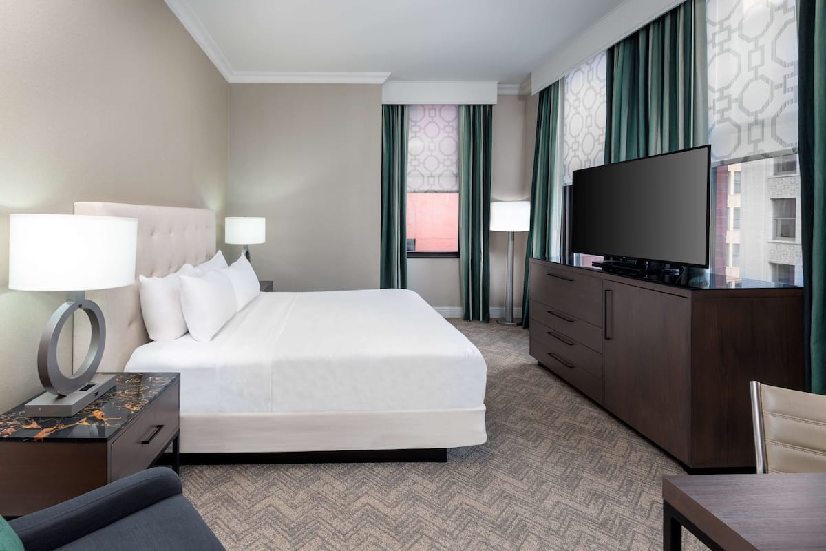A hotel room with a large bed, a TV, a desk with a lamp, and a view of buildings through the window.