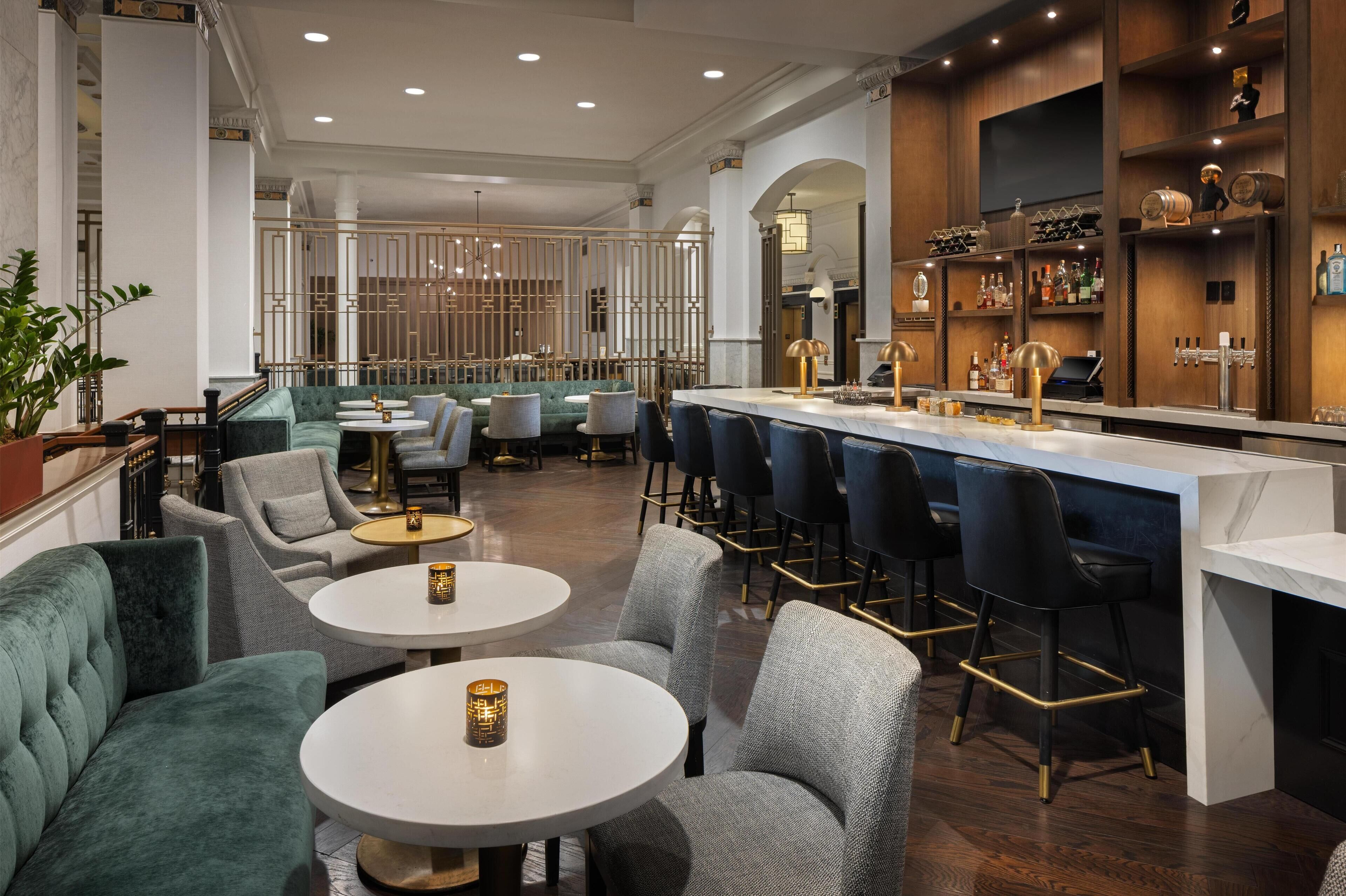 A bar area with a marble countertop, bar stools, and a variety of bottles and glasses. There are round tables with chairs and a seating area with sofas.