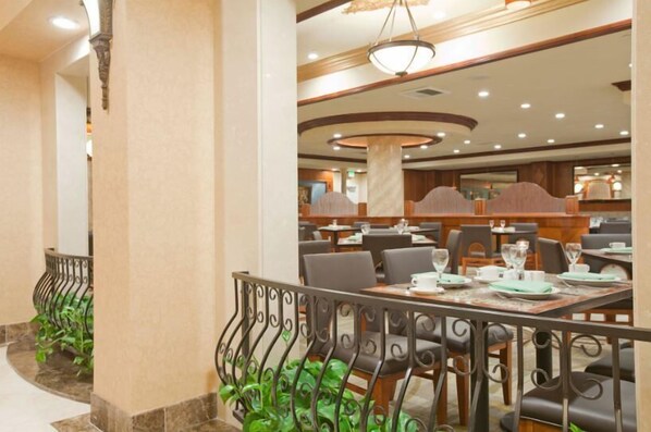 A dining area with tables set for service, a curved ceiling with recessed lighting, and a decorative railing with greenery.