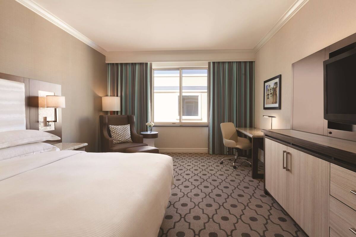 Une chambre d’hôtel avec un grand lit, un bureau, une chaise, une télévision et une fenêtre avec des rideaux.