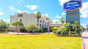 Edifici d'un Hampton Inn & Suites by Hilton, amb un rètol, envoltat d'un jardí ben cuidat i palmeres.