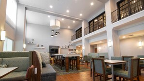 Un hall d’hôtel spacieux avec un plafond haut, plusieurs espaces de détente comprenant un canapé et des fauteuils, une table à manger centrale et un escalier menant à un étage supérieur.