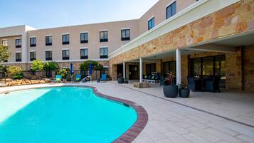 A swimming pool with a brick edge, surrounded by a covered patio area with outdoor seating and potted plants.
