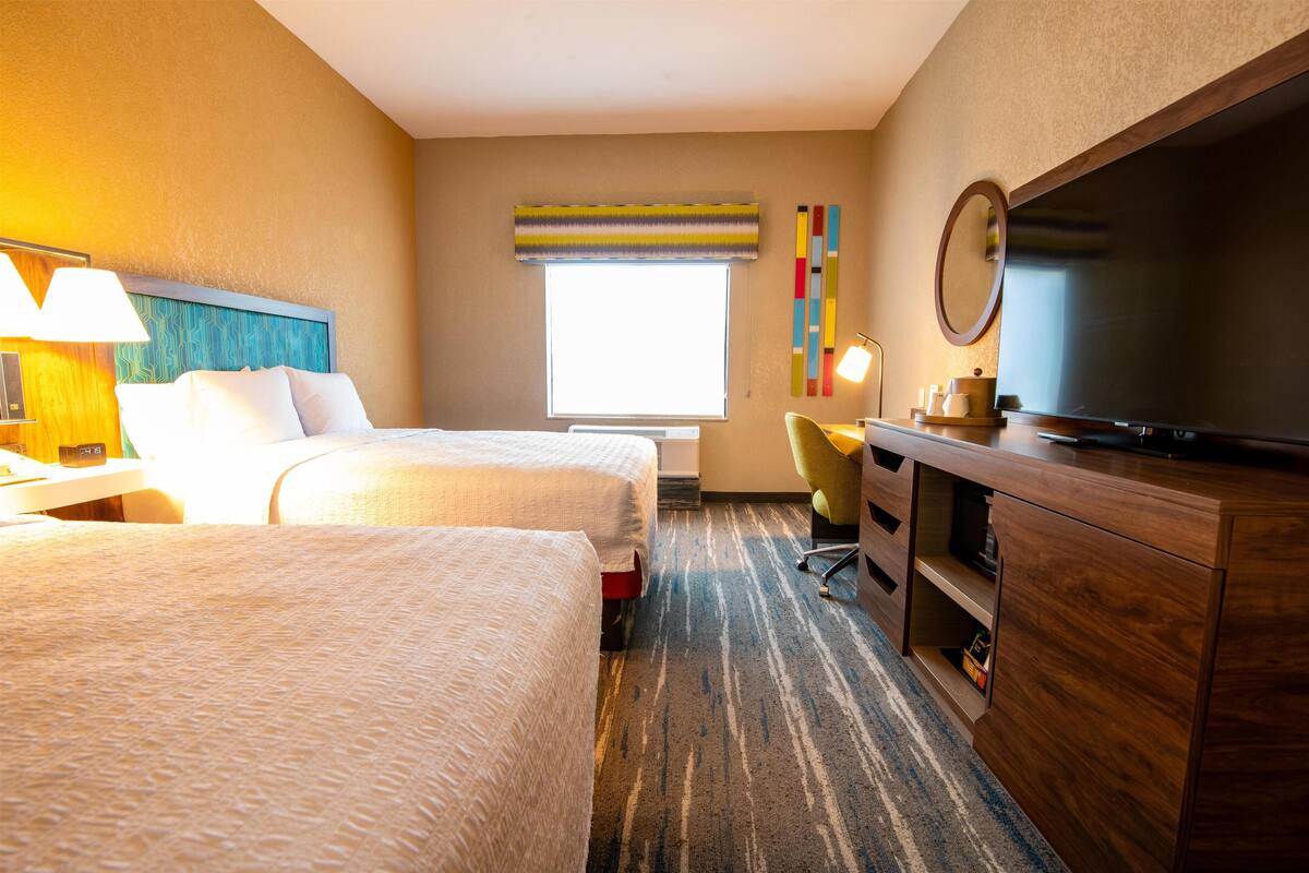 A hotel room with two beds, a desk with a lamp, a TV, a chair, and a large window with a view.