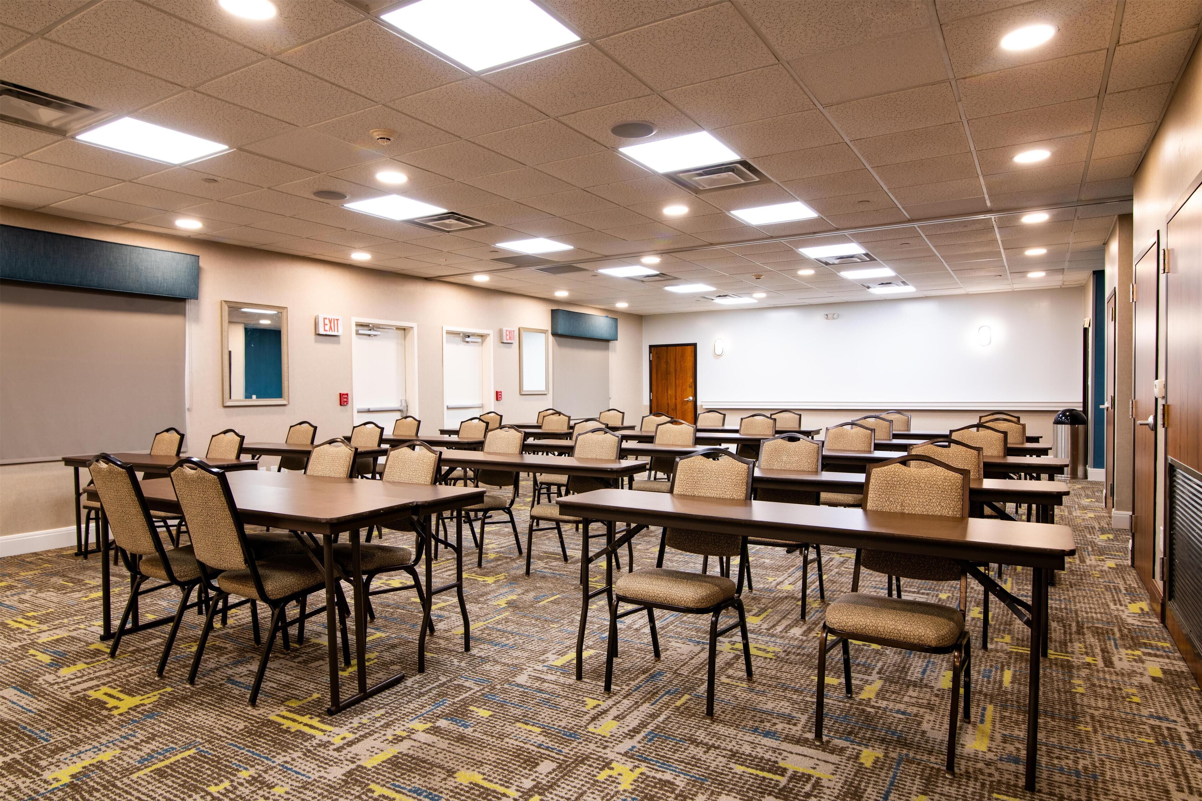 A conference room with multiple tables and chairs, a projector screen, and an exit sign.