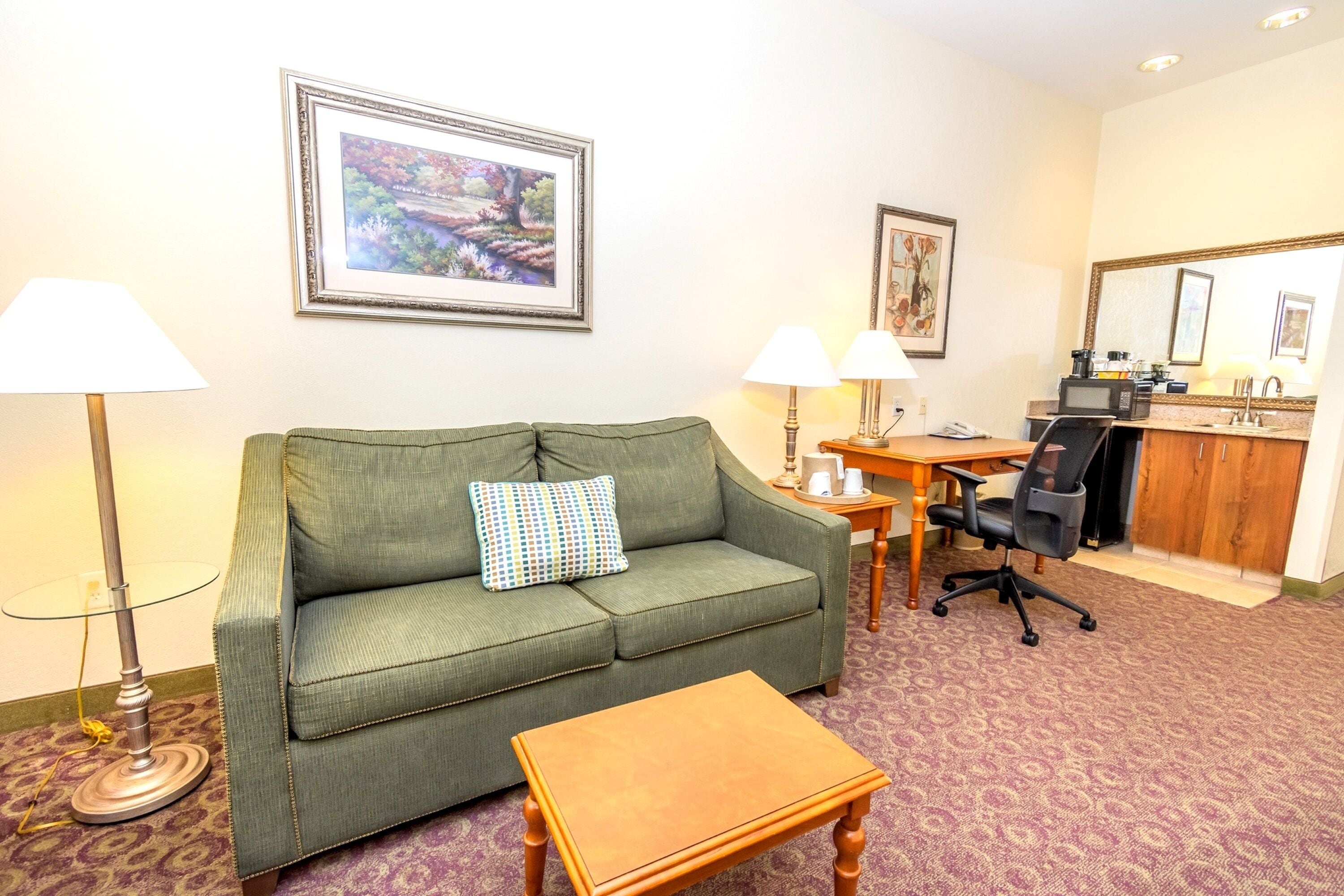 Quarto de hotel com sofá verde, mesa de centro de madeira, escrivaninha com cadeira, micro-ondas e pia.