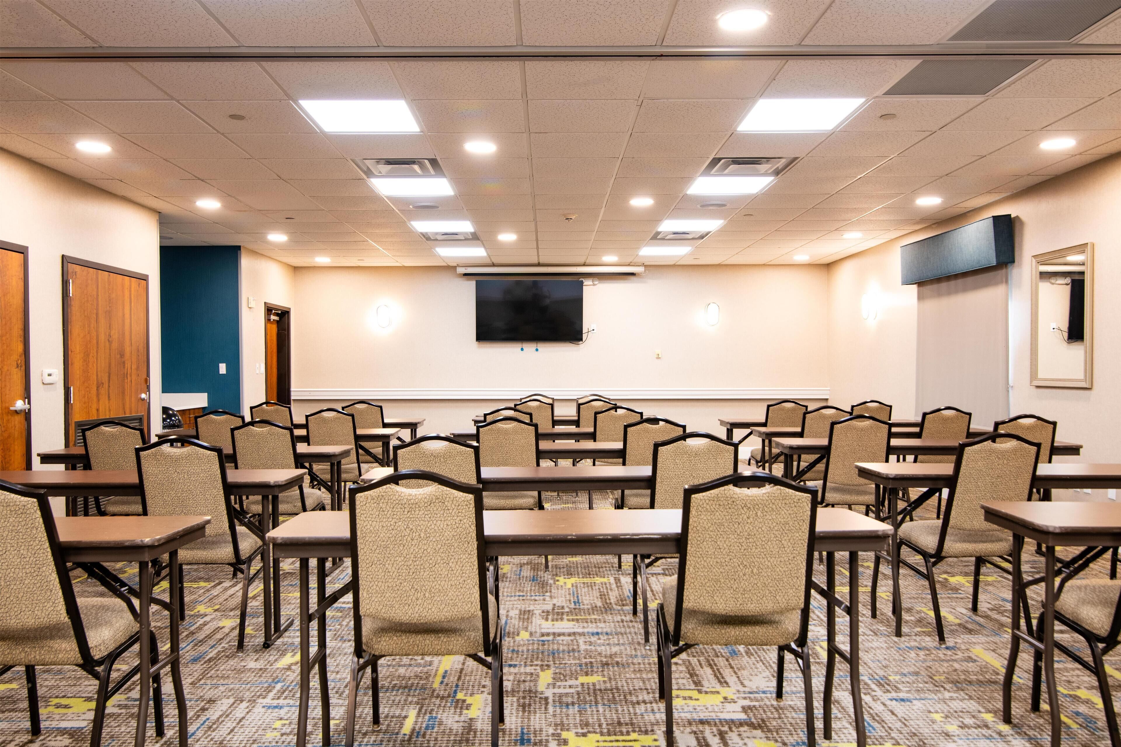 A conference room with multiple tables and chairs, a flat-screen TV on the wall, and a carpeted floor.