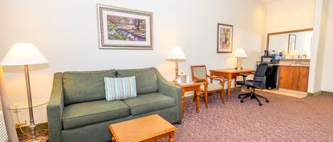 A hotel room with a green sofa, a wooden coffee table, a desk with a chair, and two lamps.