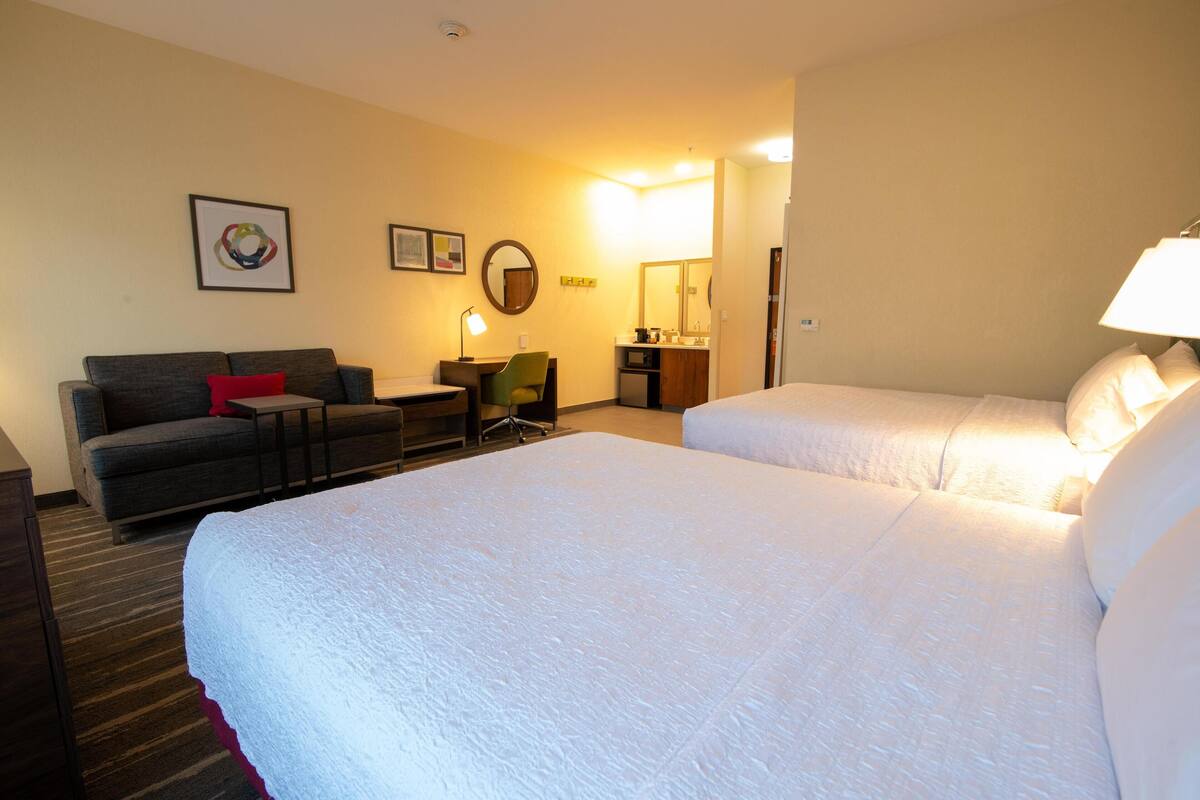 A hotel room with two beds, a desk, a chair, a sofa, a lamp, and a mirror.