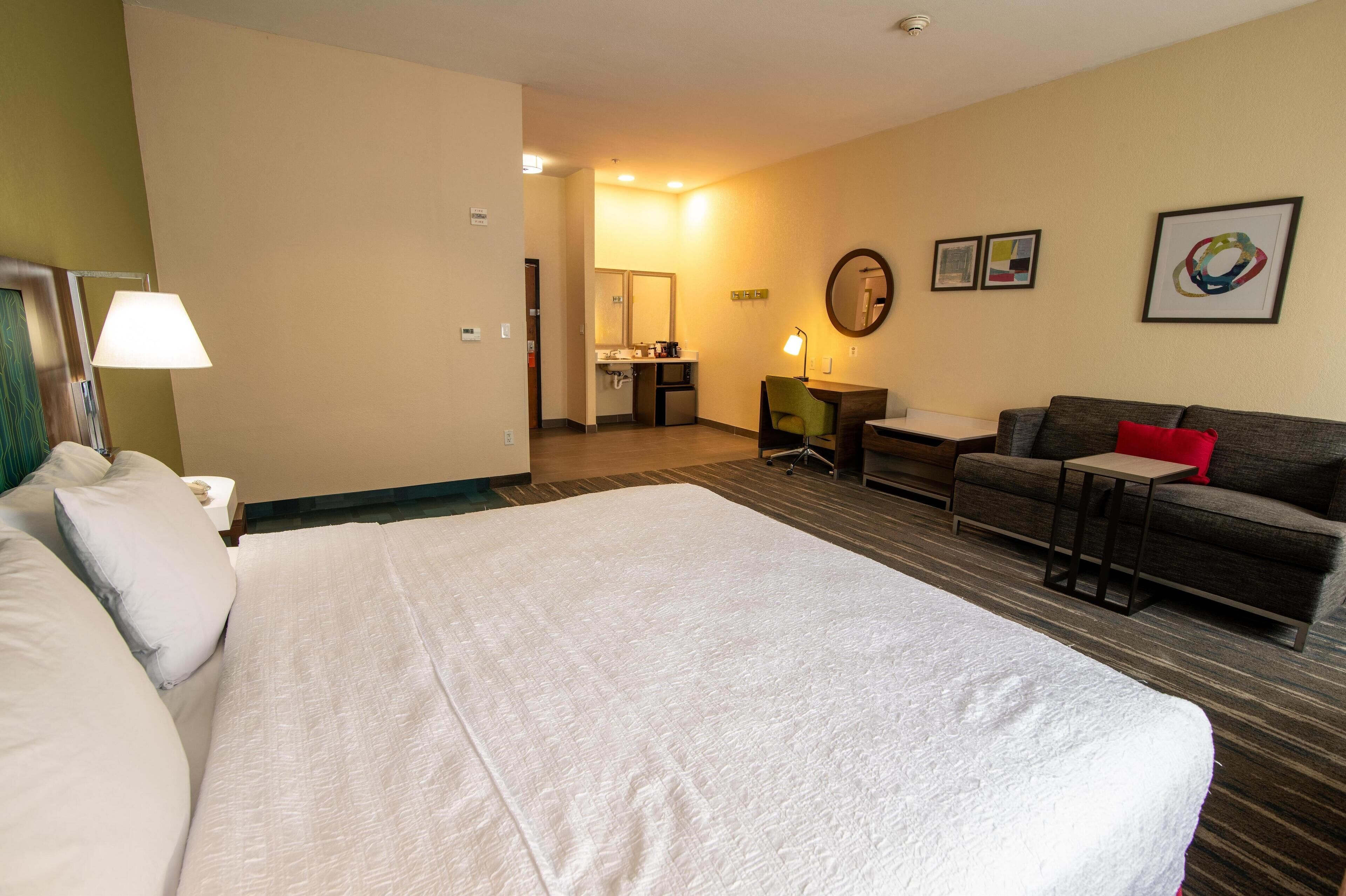 A hotel room with a large bed, a sofa, a desk, and a small table. There is a bathroom visible in the background.