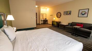 A hotel room with a large bed, a sofa, a desk, and a small table. There is a bathroom visible in the background.