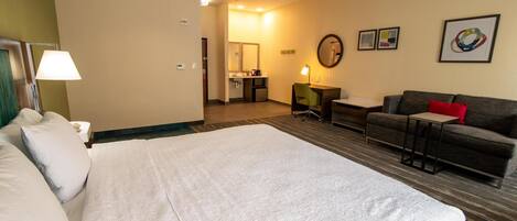 A hotel room with a large bed, a sofa, a desk, and a small table. There is a bathroom visible in the background.