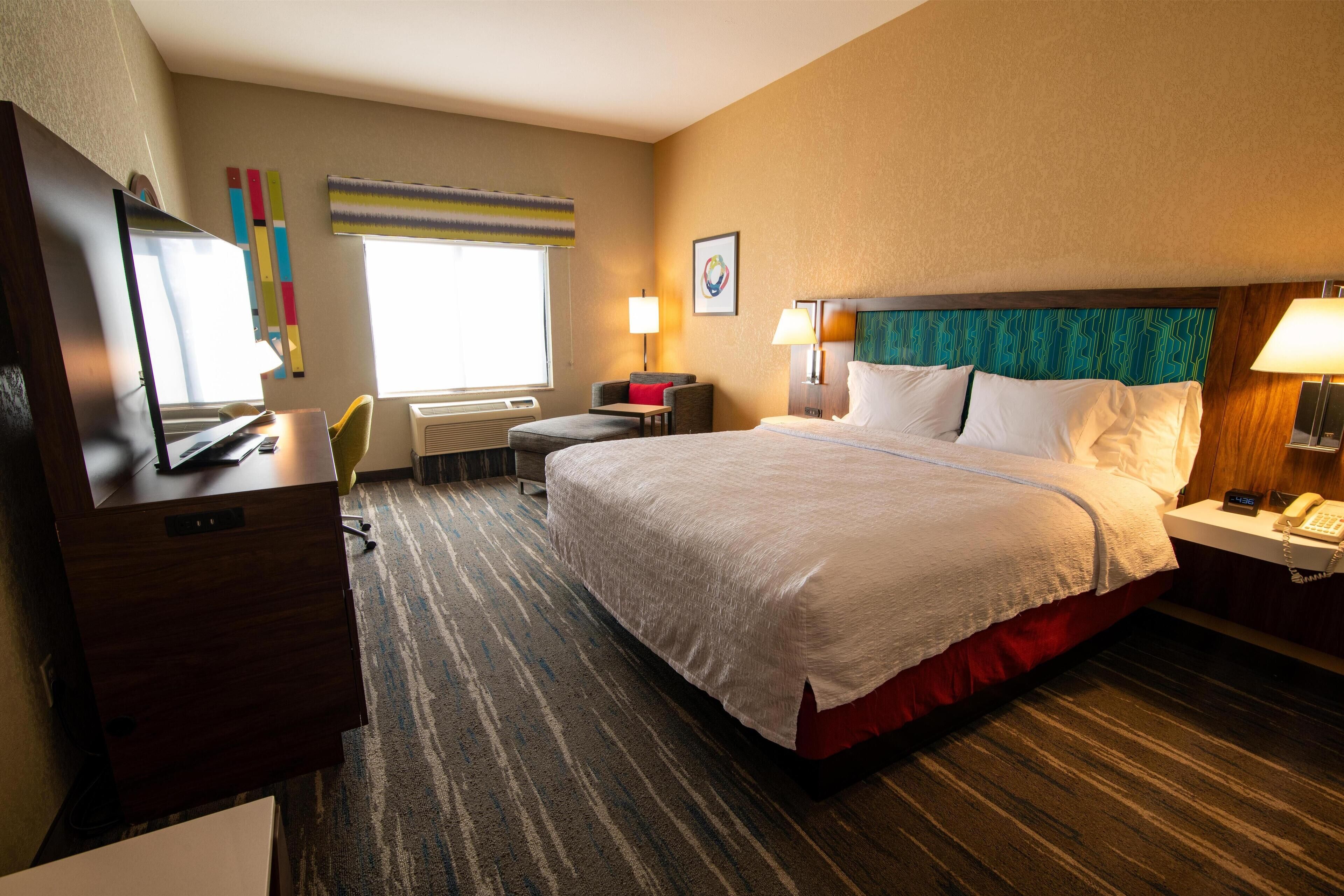 Habitación de hotel con una cama grande, un escritorio con computadora, una silla, una ventana con persianas y una pared con una obra de arte colorida.