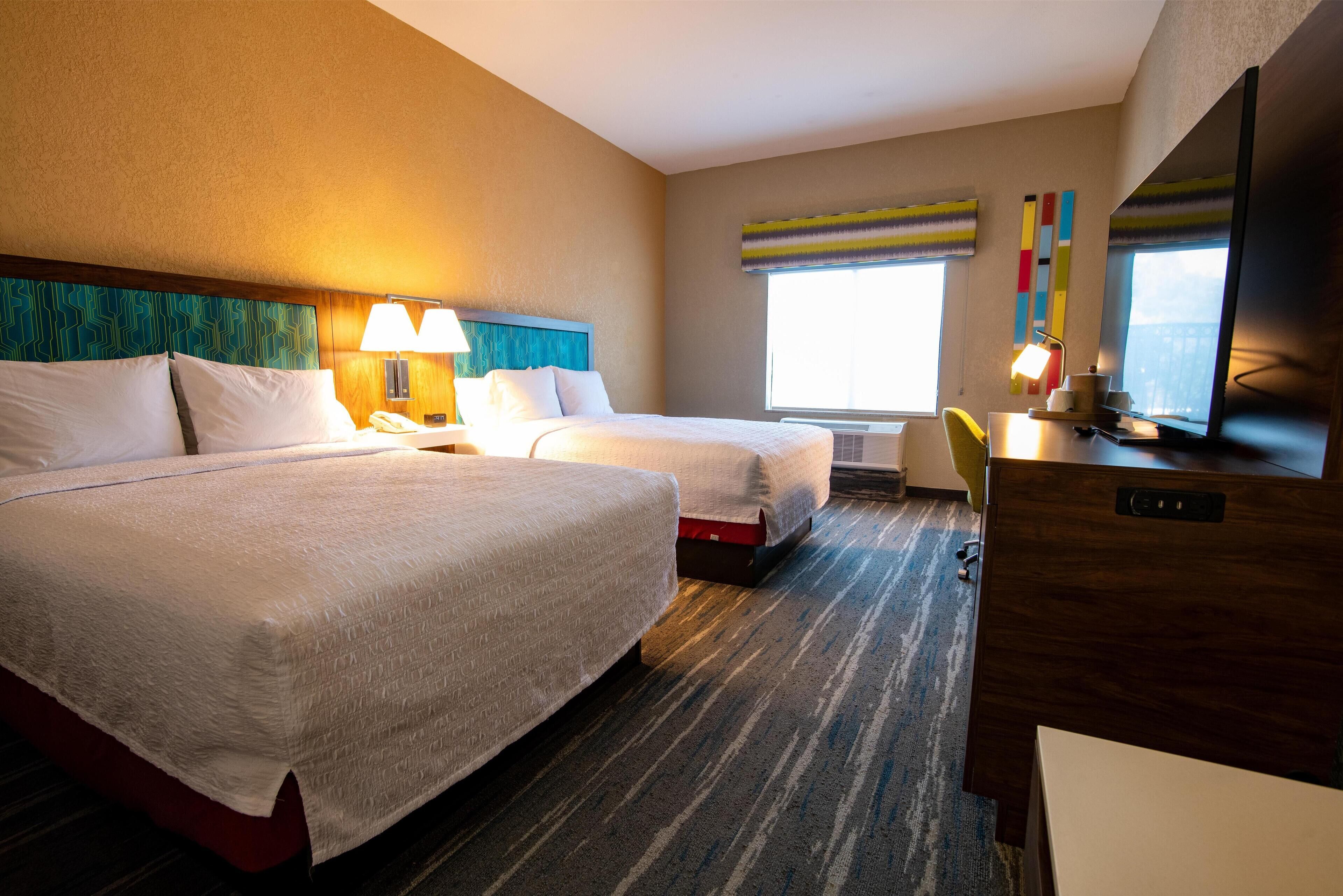 A hotel room with two beds, a desk, a chair, a TV, and a window with blinds.