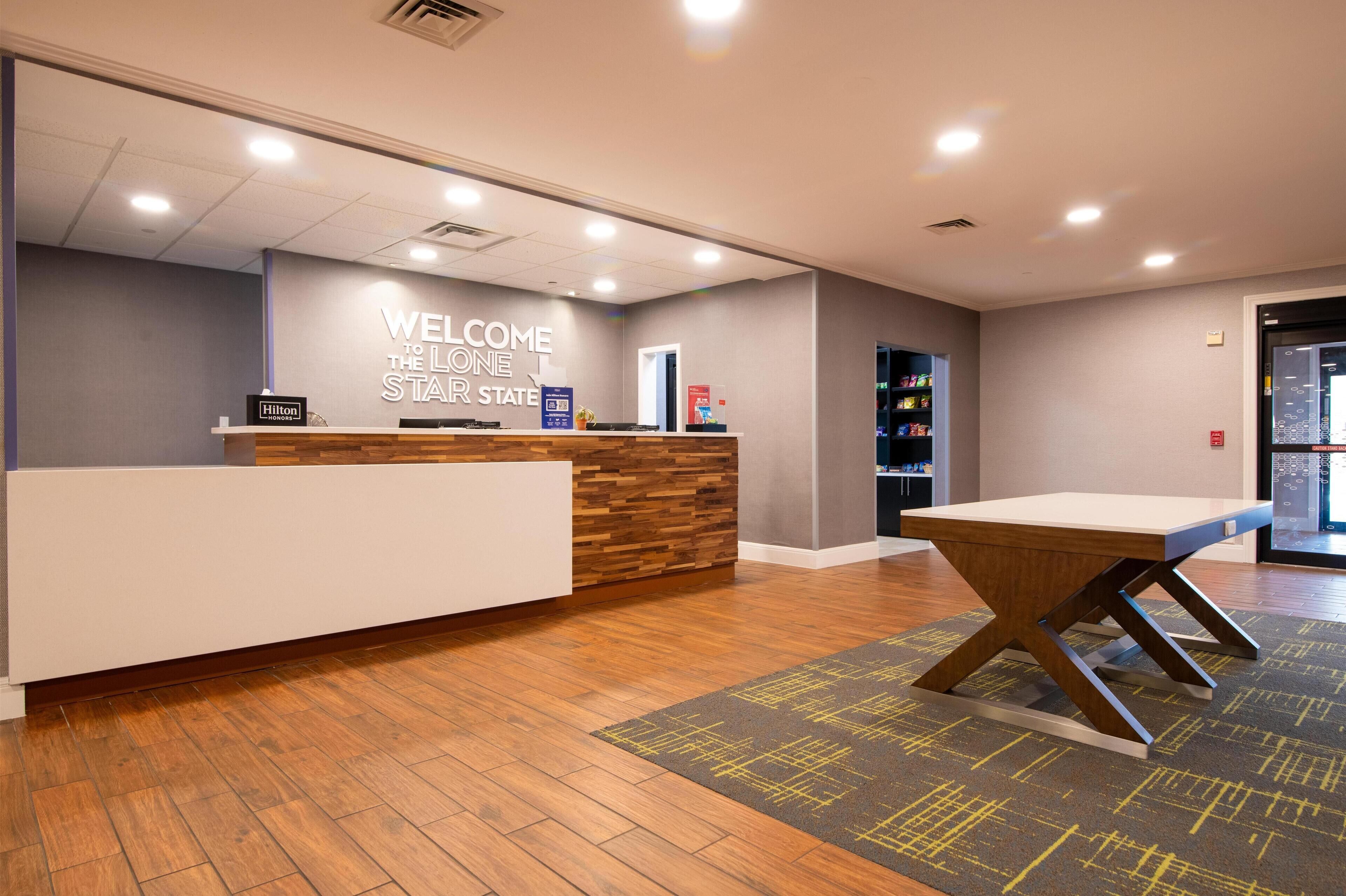 En hotellobby med en receptionsdisk i trä, en skylt med texten ”Välkommen till Lone Star State” och en modern design med trägolv och ett geometriskt mönster i mattan.