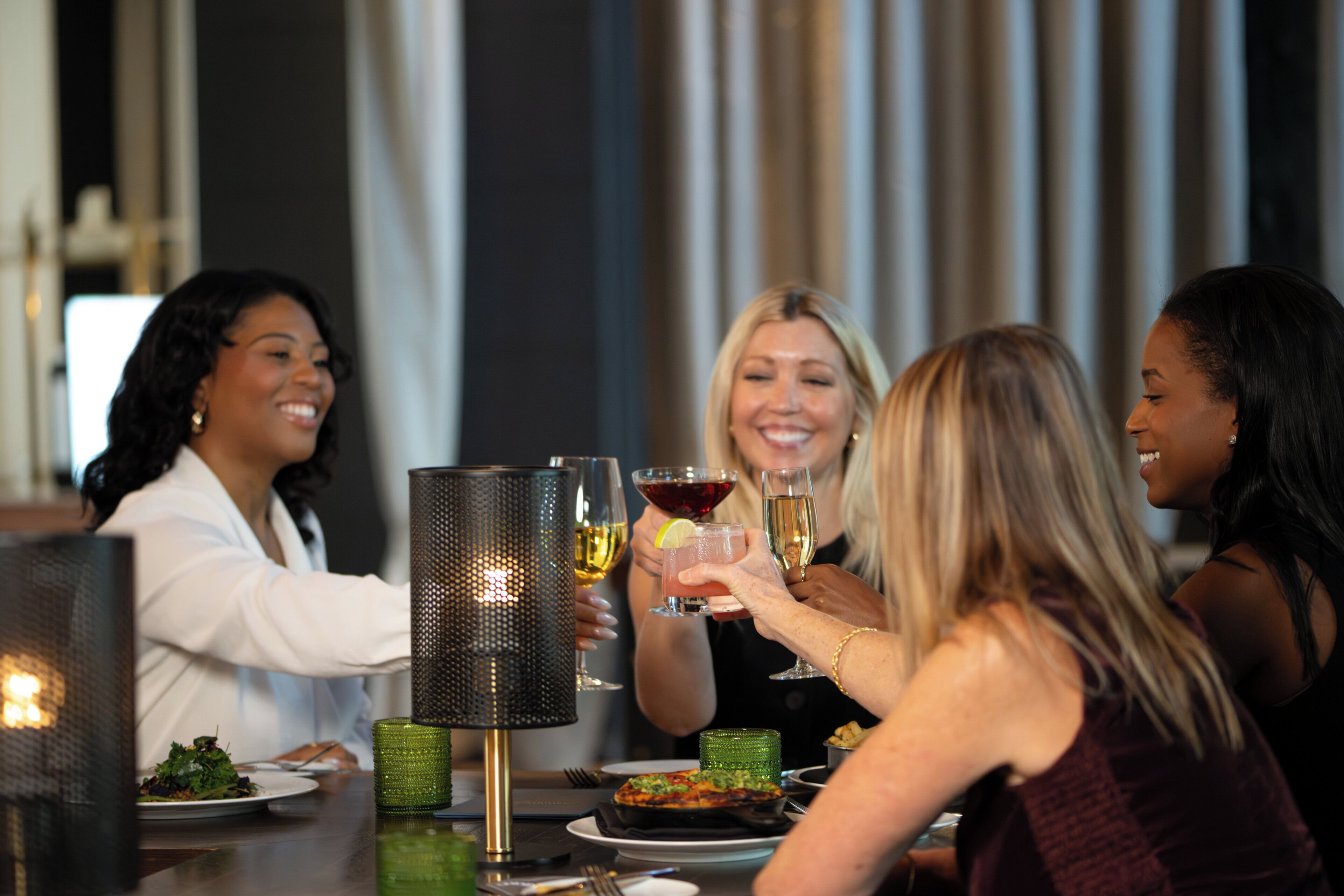 Un grupo de personas brindando con bebidas en una mesa con una lámpara decorativa, platos de comida y una vela.