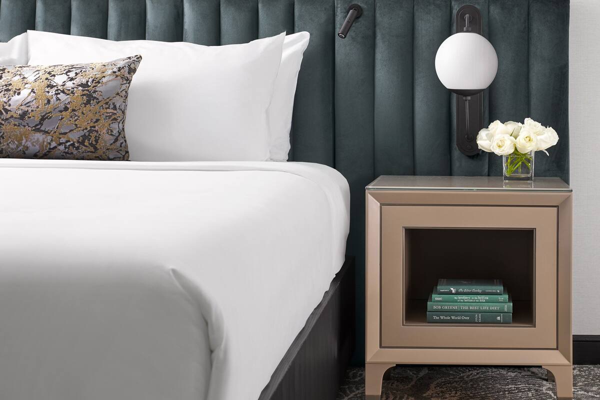 A neatly made bed with white linens and a decorative pillow. A small bedside table with a lamp and a vase of flowers is beside the bed.