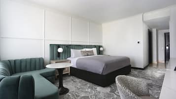 A modern hotel room with a sectional sofa, a round table, a bed with a headboard, and a patterned carpet.