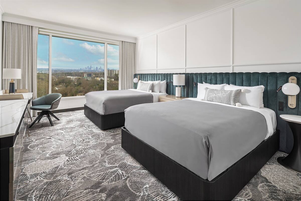 A hotel room with two beds, a desk, a chair, and a large window offering a city view.
