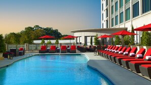 Un área junto a la piscina con sillas de descanso y sombrillas rojas, una piscina azul cristalina y un edificio con grandes ventanales al fondo.