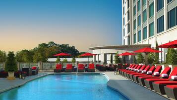 A swimming pool with lounge chairs and red umbrellas, surrounded by a building with large windows.