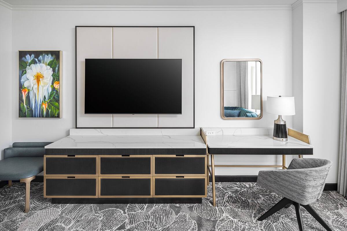 A modern hotel room with a large flat-screen TV, a sofa, a desk with a lamp, and a chair.