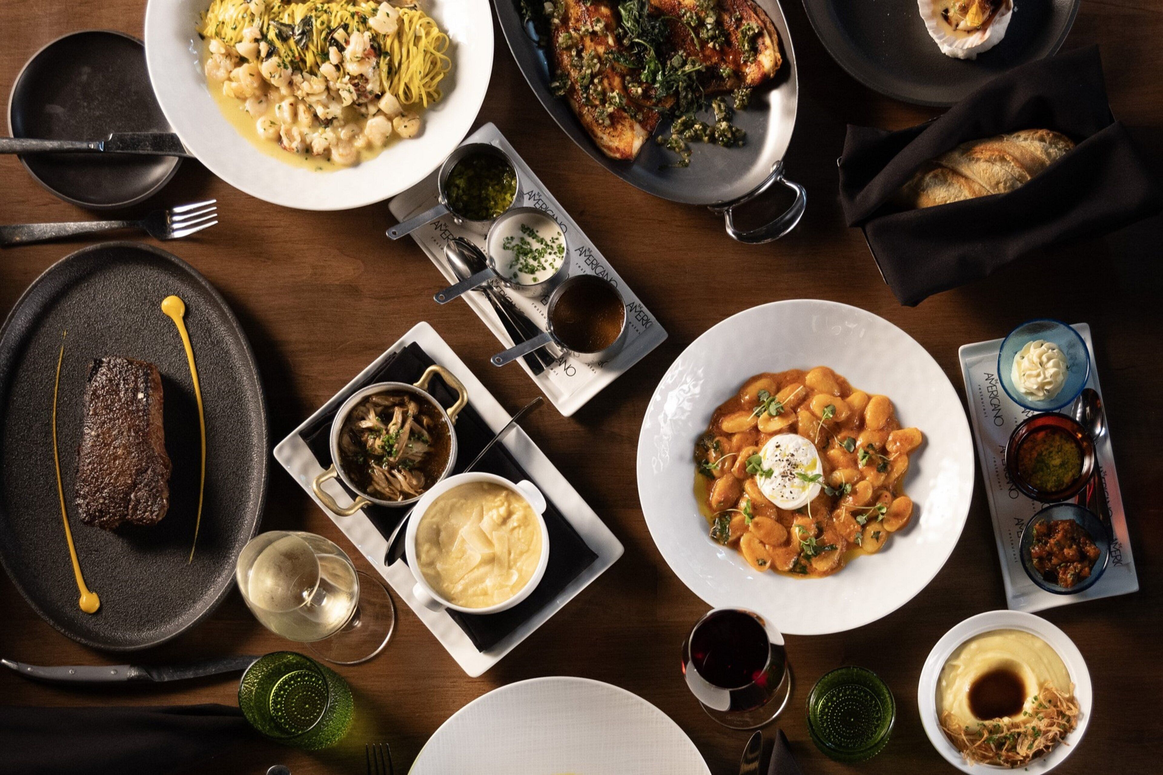A table set with various dishes, including pasta, steak, and bread, along with multiple glasses of wine.