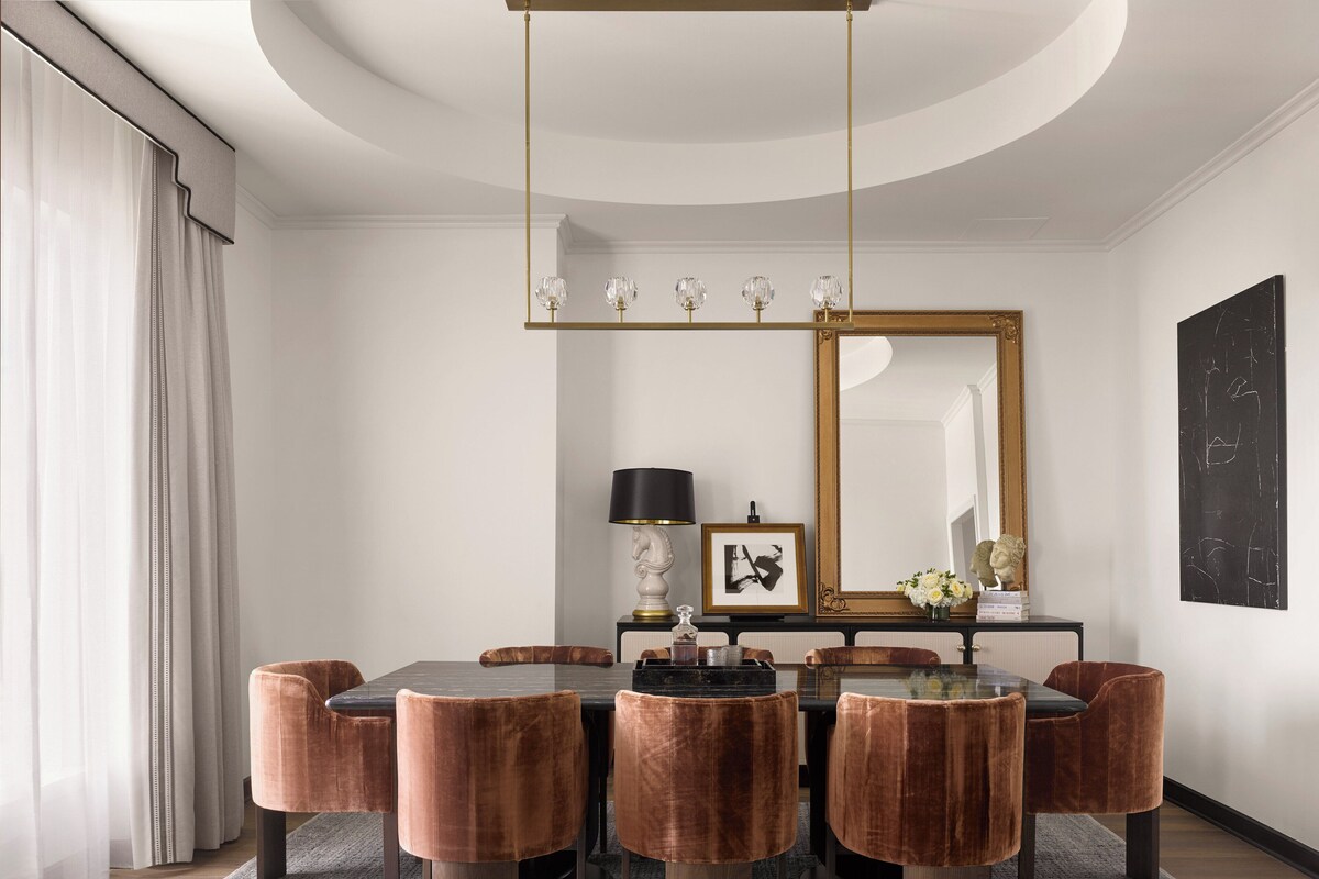 A modern dining room with a round table, six velvet chairs, a chandelier, a large mirror, and a framed picture.