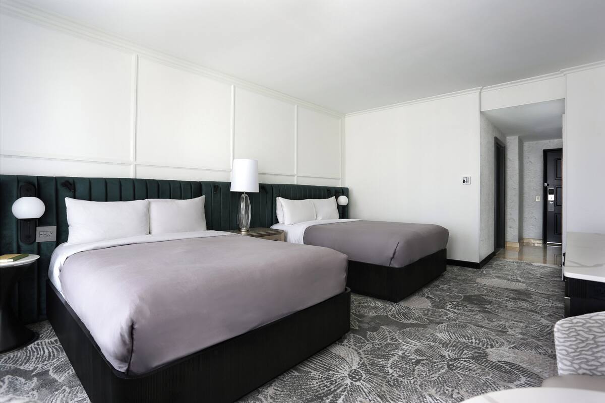 A hotel room with two beds, a nightstand with a lamp, a desk, and a patterned carpet.