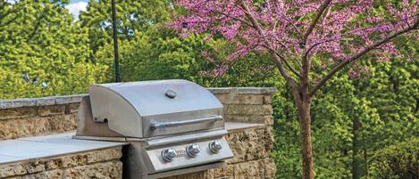 A stone barbecue grill with a control panel, set against a backdrop of trees and a clear sky.