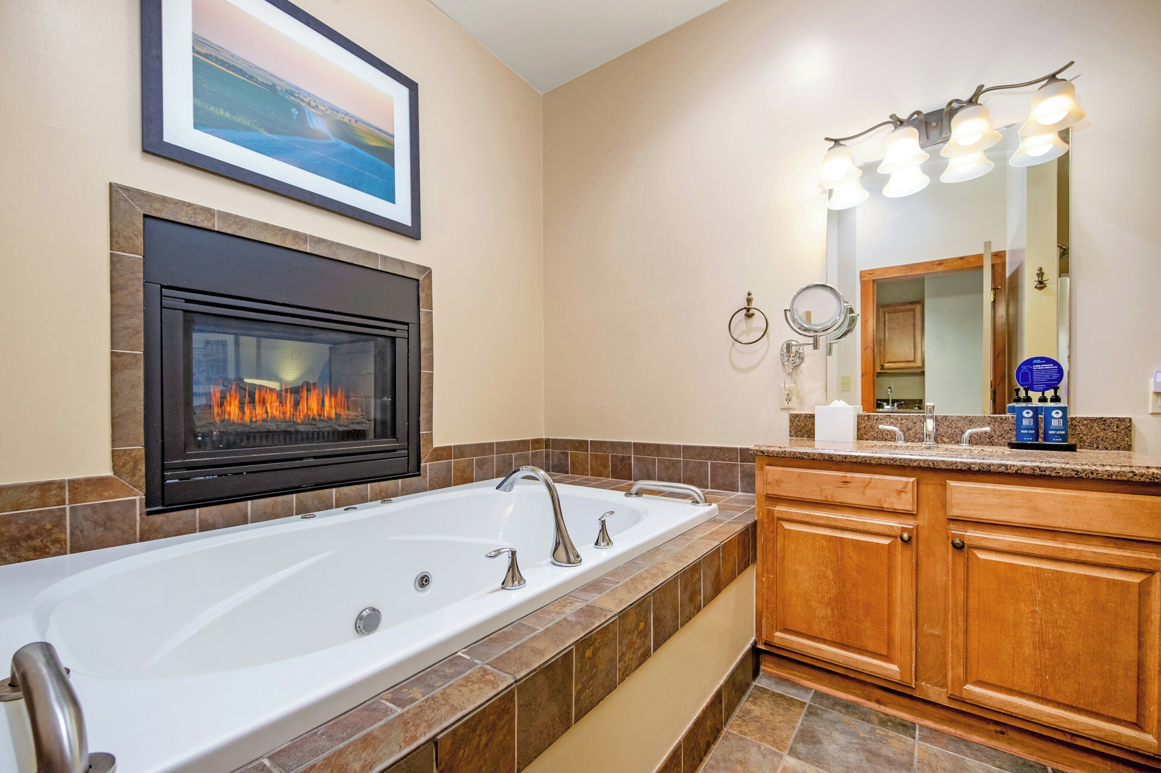 A bathroom with a large bathtub, a fireplace, a mirror with lights, and a framed picture on the wall.