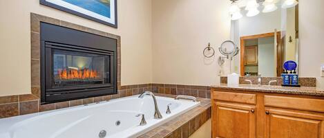 A bathroom with a large bathtub, a fireplace, a mirror with lights, and a framed picture on the wall.