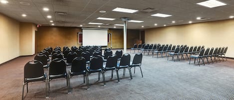 Egy konferenciaterem, melyben sorokban állnak a székek, van egy vetítővászon és a padló szőnyeggel van borítva.
