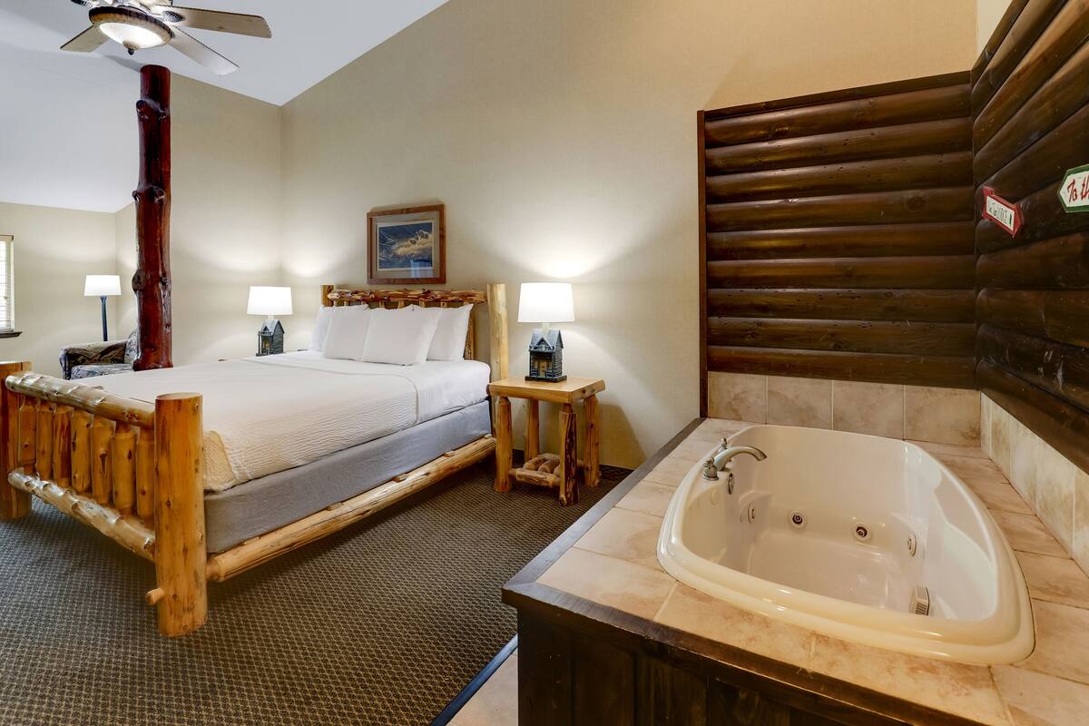 A bedroom with a wooden bed, two bedside lamps, a ceiling fan, a large window, and a bathroom with a bathtub and a wooden vanity.