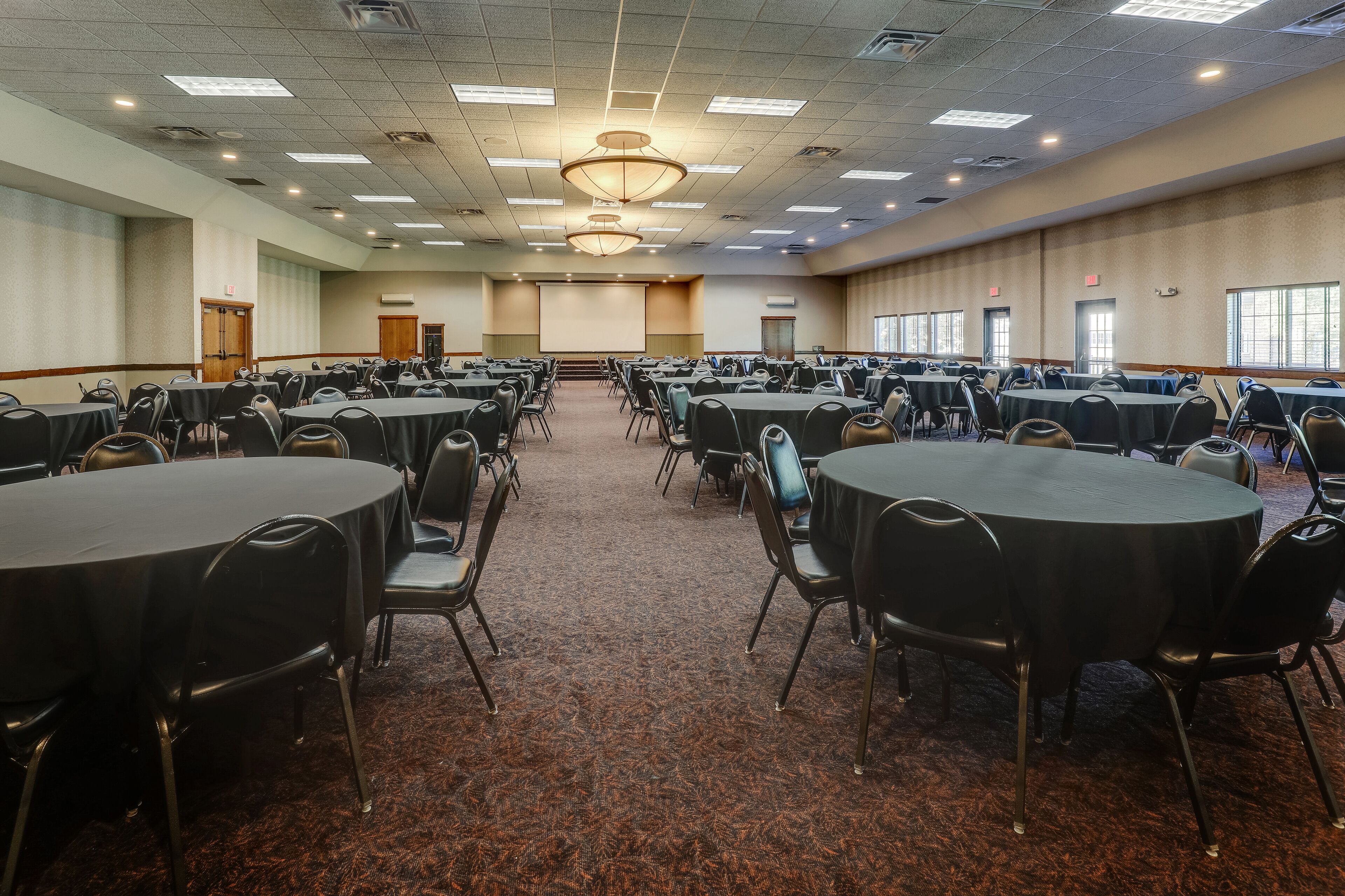 Une grande salle de conférence dotée de plusieurs tables rondes et de chaises, d’un sol recouvert de moquette et d’un écran de projection à l’avant.