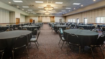 Een ruime congreszaal met meerdere ronde tafels en stoelen, een vloerbedekking en een projectiescherm aan de voorzijde.