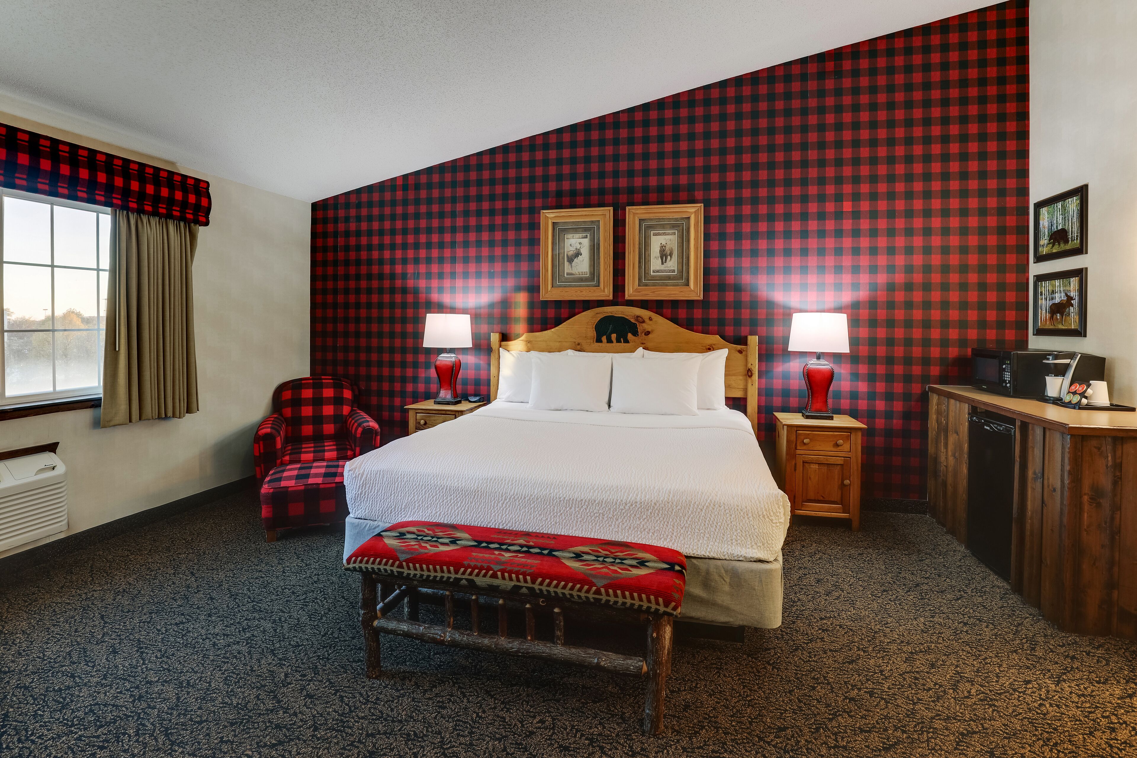 A hotel room with a large bed, two bedside tables with lamps, a wooden bench, a red plaid chair, and a checkered wall.