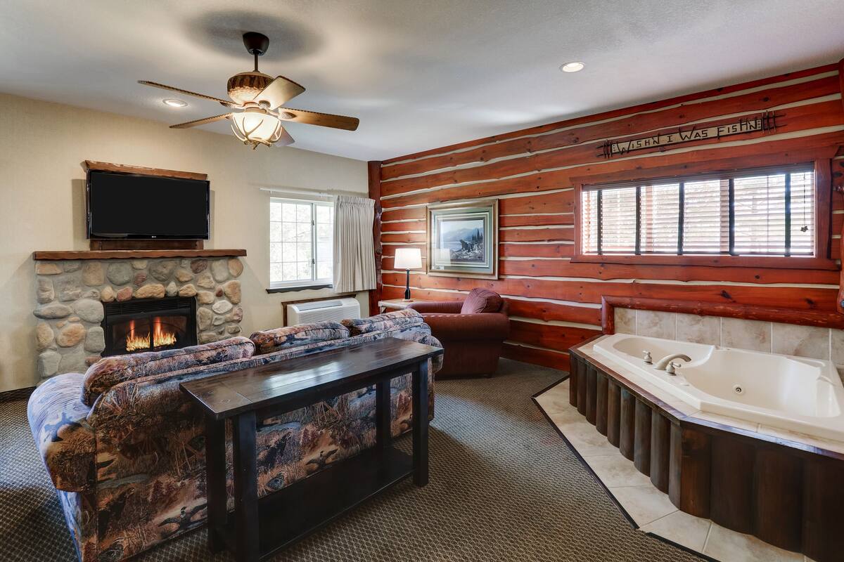 A log cabin interior with a stone fireplace, a large sofa, a television, a ceiling fan, a window with blinds, a framed picture, and a bathtub.