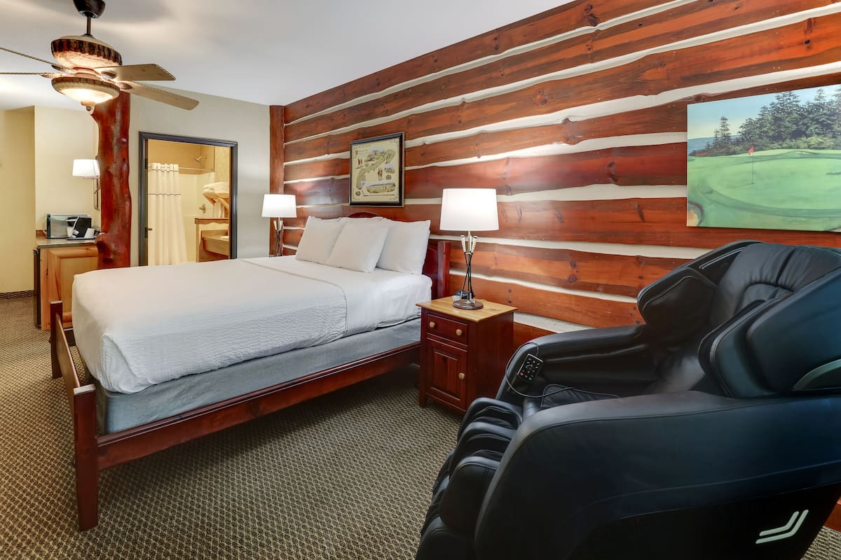 A hotel room with a wooden wall, a bed, a massage chair, and a painting of a golf course.